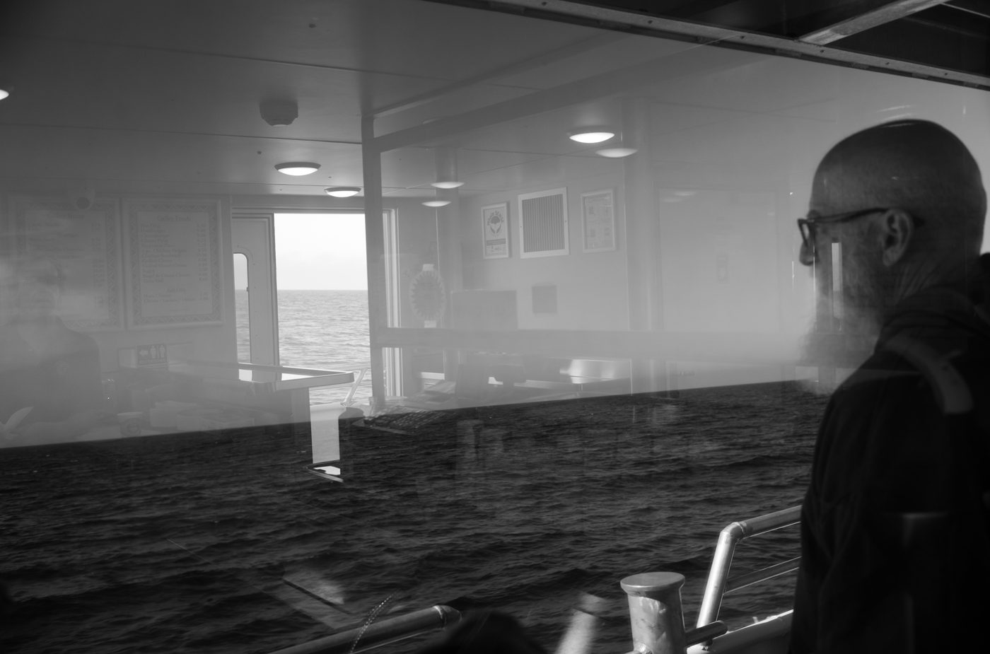 A passenger on a ship looks out towards the ocean seen through the reflection in the glass behind him.