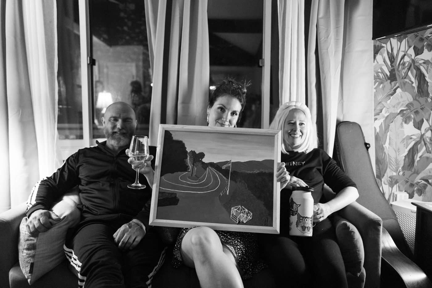 A woman sitting on a couch holds a painting up to the camera
