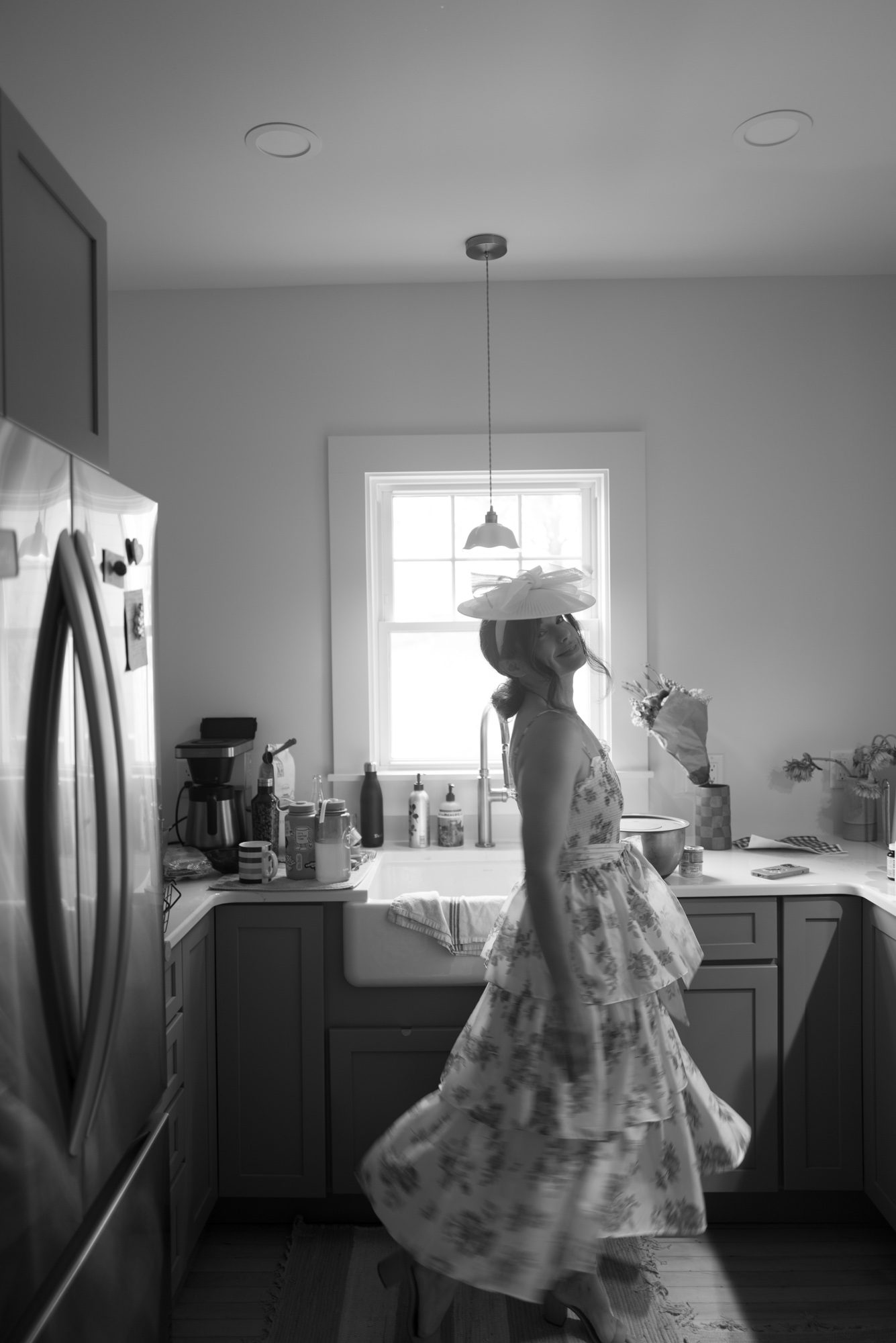 A woman in a fascinator turns from a kitchen sink to face the camera 